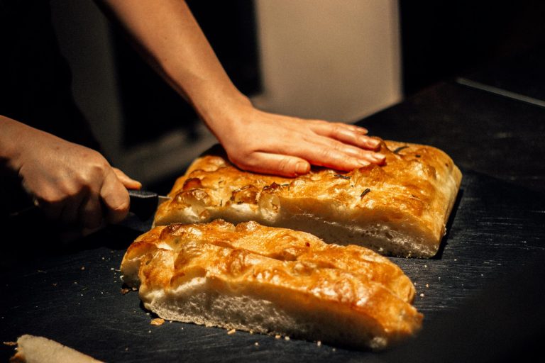 Close-up of hands cutting freshly baked focaccia bread on a dark surface.
