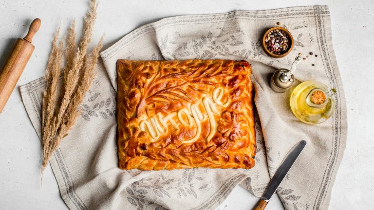 Golden brown homemade pie with decorative crust on linen fabric surrounded by spices and oil.